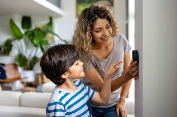 A mother and child smile while adjusting a smart thermostat on the wall inside their home.